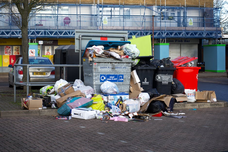 A collection of overflowing waste and rubbish bags piled around two large wheeled bins on a paved street in front of commercial stores, including a paper and cardboard recycling bin and a general waste bin. The rubbish includes cardboard boxes, plastic bags, paper, and packaging materials, some spilling onto the ground. A metallic railing separates the waste area from a parked silver car with a yellow license plate. In the background, there is a building under construction or renovation with scaffolding and protective netting, and storefront signs indicating various shops and services. The scene appears typical of private waste accumulation for disposal or collection, highlighting the importance of effective rubbish removal services in urban environments.