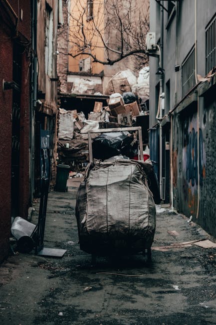A narrow alleyway cluttered with large piles of mixed waste materials, including black and white plastic bags, cardboard boxes, and loose debris, stacked against a wall at the far end. The foreground features a sizable, worn, grey fabric sack or bag on wheels, positioned centrally, with visible creases and dirt marks. The surrounding environment includes building facades on either side, with one side featuring graffiti and metal grilles, while the other shows exposed brick and some wiring. Bare tree branches extend over the alley overhead, and the lighting appears subdued, suggesting an overcast day or shaded area. The scene exemplifies typical private waste collection or clearance activity, with Waste Removal Hammersmith potentially handling removal of household or commercial rubbish in such urban alleyways, emphasizing the need for effective rubbish management in confined spaces.