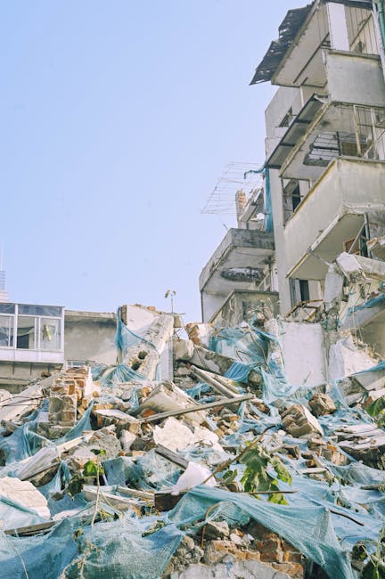 A narrow alleyway cluttered with large piles of mixed waste materials, including black and white plastic bags, cardboard boxes, and loose debris, stacked against a wall at the far end. The foreground features a sizable, worn, grey fabric sack or bag on wheels, positioned centrally, with visible creases and dirt marks. The surrounding environment includes building facades on either side, with one side featuring graffiti and metal grilles, while the other shows exposed brick and some wiring. Bare tree branches extend over the alley overhead, and the lighting appears subdued, suggesting an overcast day or shaded area. The scene exemplifies typical private waste collection or clearance activity, with Waste Removal Hammersmith potentially handling removal of household or commercial rubbish in such urban alleyways, emphasizing the need for effective rubbish management in confined spaces.