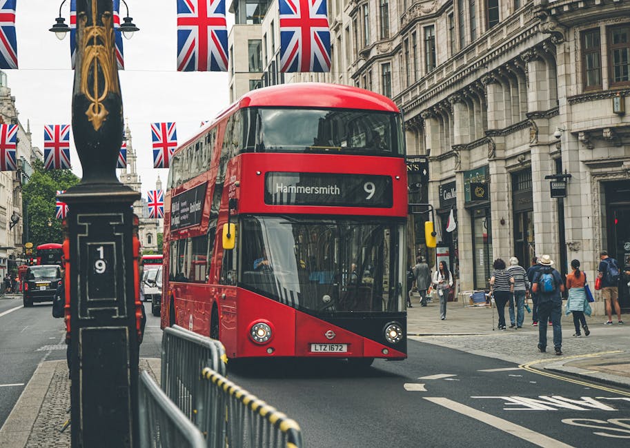 A red double-decker bus displaying the route to Hammersmith, positioned in the middle of a busy urban street with historic buildings and storefronts on either side. The bus is modern, with a sleek design and large front windows, and is flanked by pedestrians walking on the sidewalk. Some individuals are carrying shopping bags or backpacks, dressed in casual clothing suitable for a city environment. The street is lined with decorative lampposts, and the front of a black lamppost with gold embellishments is visible in the foreground. Numerous Union Jack flags hang overhead across the street, adding a decorative touch to the scene. To the left of the bus, a metal barrier separates the road from the sidewalk, and a small black roadside object with a white number '19' is also present. The lighting is natural, with overcast skies casting a diffuse light over the scene, illustrating typical city traffic and pedestrian activity, relevant to urban transport and private travel options in Hammersmith.