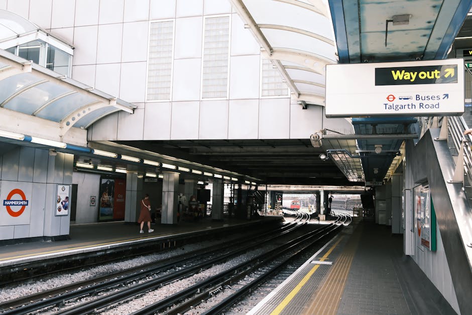 The image displays a section of Hammersmith underground station platform with multiple train tracks running through the middle, bordered by raised concrete and tiled surfaces with yellow tactile paving strips along the edge for safety. On the left side, a red and white Hammersmith station sign is mounted on a white tiled wall, adjacent to posters and informational signage. Several passengers are visible on the platform, some standing and others walking, dressed in casual clothing. Overhead, the station is covered by a curved, semi-transparent canopy with a metallic frame, providing shelter and natural light. To the right, a staircase with metal railings leads up from the platform, above which a black and yellow 'Way out' sign directs commuters toward an exit. The scene appears well-lit by artificial lighting, and the environment is typical of an urban rail station, with a neutral and professional atmosphere relevant to the transportation or logistical context of waste removal or material handling in an infrastructure setting.