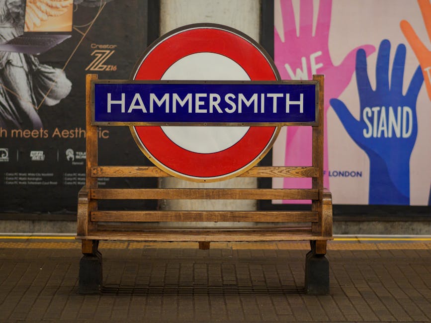 A wooden bench with a slatted seat and backrest, positioned on a paved area in front of large advertising posters in an underground station or public space. Behind the bench, there is a large round sign resembling a London Underground symbol, with a red outer circle and a white inner circle, overlaid by a blue rectangular sign with white uppercase letters spelling 'HAMMERSMITH.' To the right, part of a colorful poster displays two stylized handprints, one pink and one blue, with the words 'WE STAND' visible. The environment is illuminated with artificial lighting, casting minimal shadows, and the scene is free of clutter. This setting reflects a typical urban public transport or communal area, where the presence of the sign links to a specific district, and the materials of the bench and signage suggest durable, weather-resistant design suitable for outside or transit environments. Waste removal services like Waste Removal Hammersmith often operate in such locations for the clearance of bulky waste and rubbish in public spaces, supporting alternative disposal methods outside local authority arrangements.