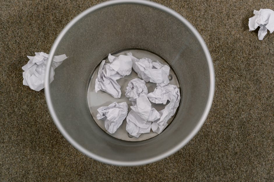 An overhead view of a cylindrical metal waste bin placed on a brown, textured carpeted flooring. The bin contains several crumpled pieces of white paper, some of which have been partially submerged in a small amount of water at the bottom. Aside from the bin, a few additional crumpled papers are scattered on the surrounding carpet, indicating a recent or ongoing tidying or rubbish removal process. The metal surface of the bin has a smooth, matte finish, reflecting minimal light, and the crumpled papers exhibit irregular folds and creases, presenting a typical paper waste scene. The environment appears to be an indoor space, likely an office or similar setting where discarding paper waste is common, aligning with private waste handling or alternative rubbish collection services. The scene emphasizes the importance of proper waste disposal in a professional context, consistent with services provided by Waste Removal Hammersmith for efficient rubbish collection and on-site clearance.