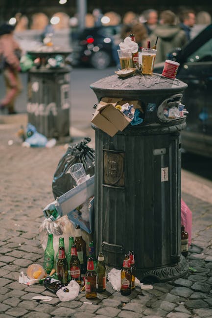 A black, cylindrical public trash bin with a domed lid situated on a cobblestone street. The bin is overflowing with various rubbish, including crumpled paper cups, plastic bottles, and discarded fast-food containers, some of which are resting on top of the lid. Several glass bottles are stacked at the base of the bin, with some lying on their sides among scattered litter and a black plastic trash bag nearby. The surrounding environment appears to be an urban area, with a blurred background showing parked cars, pedestrians, and additional rubbish instances, indicative of a busy street scene. The lighting suggests an outdoor setting, possibly during late afternoon or early evening. This scene illustrates the challenges of waste management and the importance of regular rubbish collection and private disposal services, such as those provided by Waste Removal Hammersmith, especially in public spaces with high foot traffic. The overflowing bin and litter on the ground exemplify the need for efficient rubbish removal solutions to maintain cleanliness and environmental standards in urban settings.