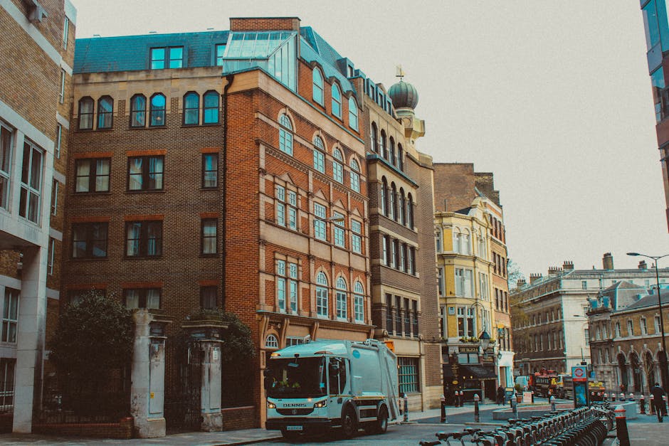 The image depicts a street scene in an urban area with a row of multi-story buildings featuring a mix of architectural styles, including brick facades, ornate window frames, and a rooftop structure with a rounded dome. In the foreground, a waste collection vehicle is parked along the curb, positioned near a gated entrance with stone pillars and greenery. The vehicle is a light-colored commercial truck with a reflective surface, indicating it is used for waste or rubbish collection services. To the right, several parked bicycles are aligned along the street, and in the distance, other vehicles, including a red double-decker bus, are visible along the road. The environment is well-lit with daylight, and the scene appears to be from a city center typical of London, with the presence of the waste removal vehicle highlighting an on-site or alternative waste handling service, such as those offered by Waste Removal Hammersmith. The overall atmosphere is quiet, with no pedestrians visible, emphasizing the urban context of rubbish collection within a city infrastructure.