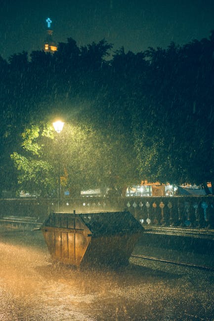 A large open-top dumpster made of weathered, rusty metal with vertical ridges is situated on a dark street curb during heavy rain at night. The dumpster is partially filled and appears to contain miscellaneous waste materials, though specific items are not identifiable. Surrounding the scene, a row of dense, leafy trees with dark green foliage line the sidewalk, illuminated faintly by nearby streetlights casting a warm yellowish glow. In the background, a wrought iron fence separates the street from a park or garden area, and a tall church steeple with a cross at its apex is visible, faintly outlined against a dimly lit sky. The rain creates small splashes as it hits the pavement and the surface of the dumpster, with the wet environment reflecting the streetlights and distant building lights. The scene captures an independent waste collection or disposal scenario, highlighting a moment of on-site rubbish storage that Waste Removal Hammersmith could service for local clients seeking private or alternative waste handling solutions.